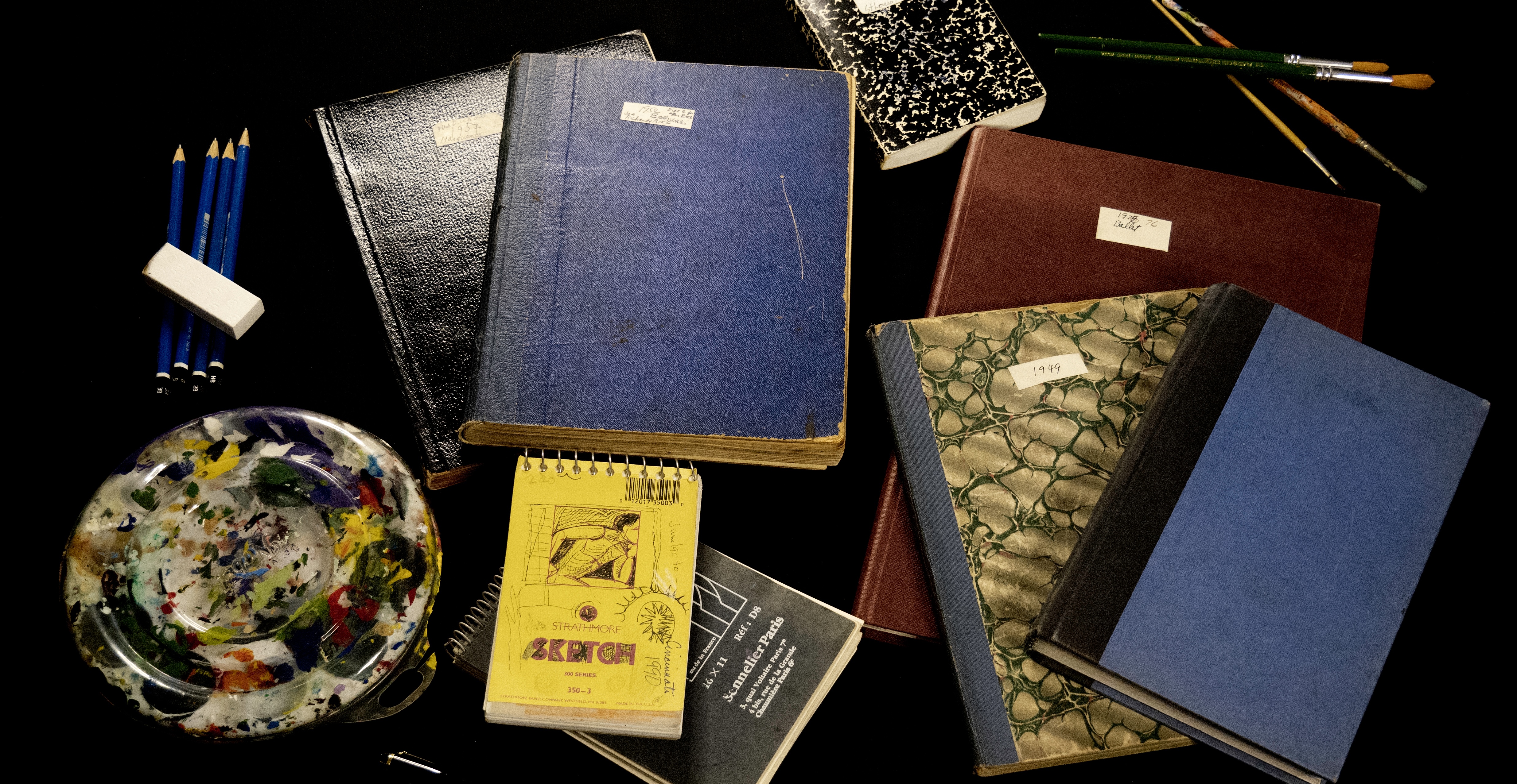 An array of closed sketchbooks in varying sizes and colours, taken from the Rita Brianksy fonds. They are strewed on a black surface along with pencils, brushes, and a painting palette. This is a screenshot from the video about Rita Briansky's sketchbooks.